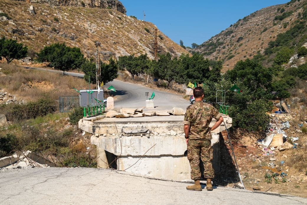 A soldier stands at a checkpoint on a winding road through arid hills, with green flags and a concrete barrier visible.