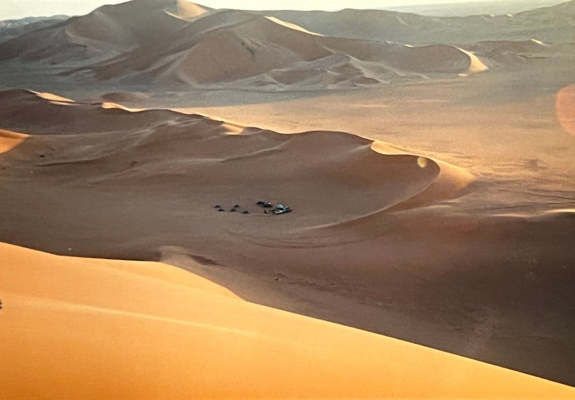 Wide aerial view of vast sand dunes at dusk with warm lighting and a small group of vehicles resting in a valley.