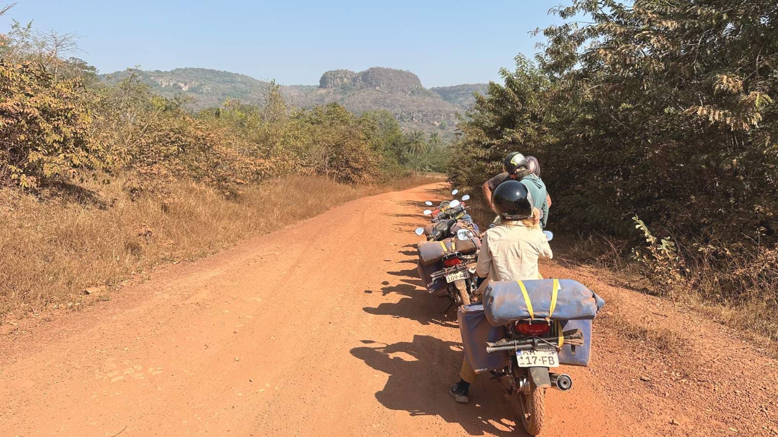 Motorcyclists with loaded panniers on a red dirt road through a hilly, green landscape under a clear sky in the Guinea highlands.