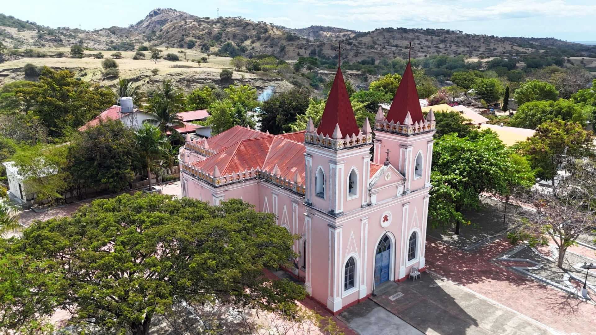 Aerial view of a pink church with red spires in a green town, with dry highland hills in the background