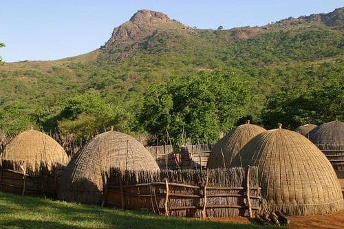 Traditional thatched dome-shaped huts enclosed by wooden fences in a rural area near Nsoko, Eswatini, set on grassy ground before green hills and a rocky peak.