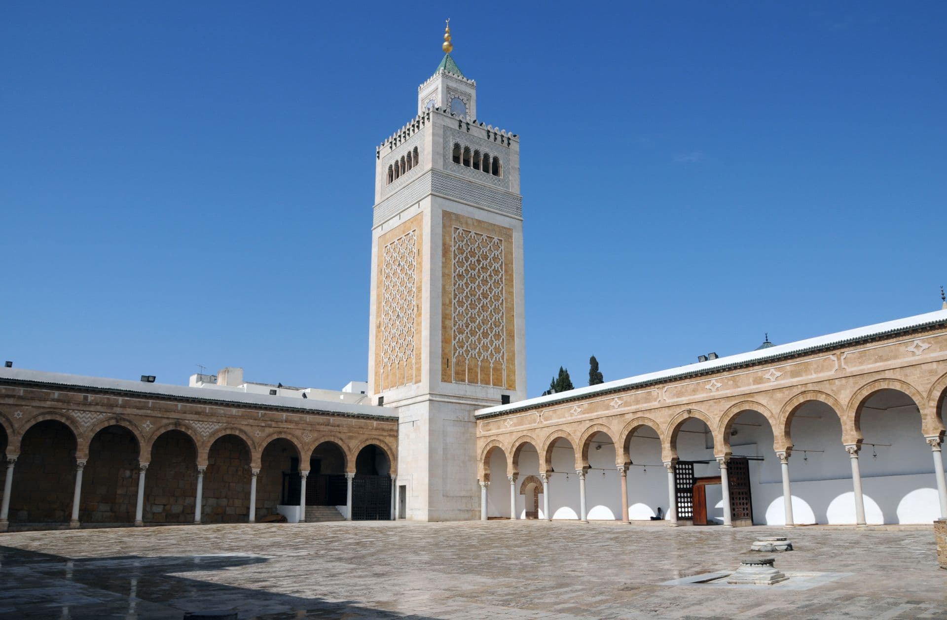 Ornate minaret with geometric tilework and arched courtyard colonnades typical of Sufi shrines in the Tunis region, under a clear blue sky.