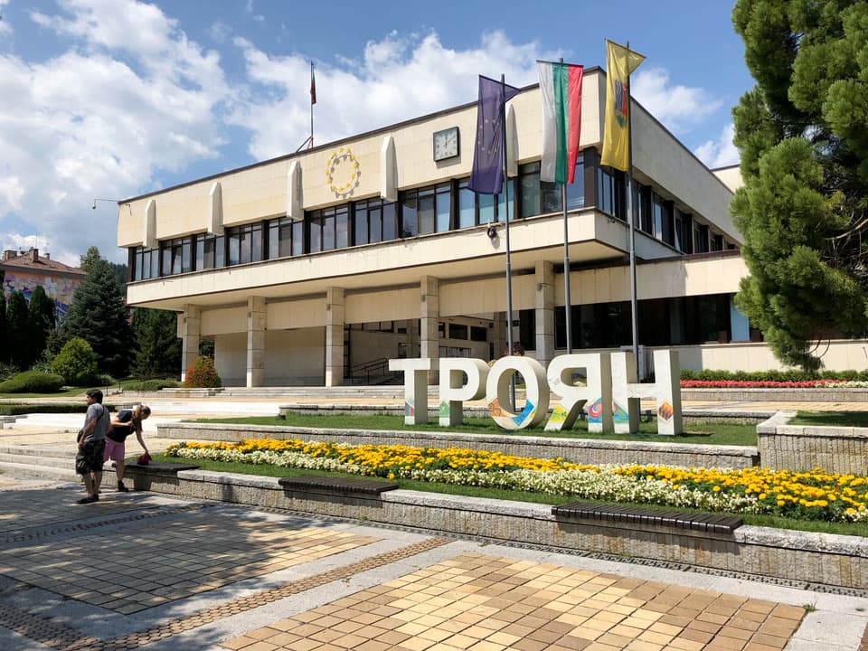 A modernist municipal building in Krichim town square features large Cyrillic signage, colorful flags, and flower beds, with two pedestrians walking on the pave
