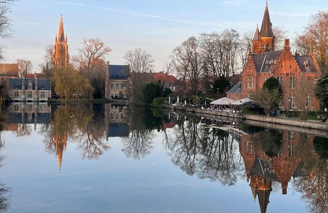 Scenic view of historic brick buildings and a church spire reflected in calm water, likely depicting Bruges rather than Bernissart.