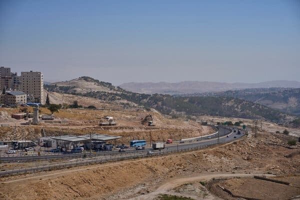 A wide highway winding through arid hills under a clear sky, with buildings and vehicles visible in the distance in the central Israel region.