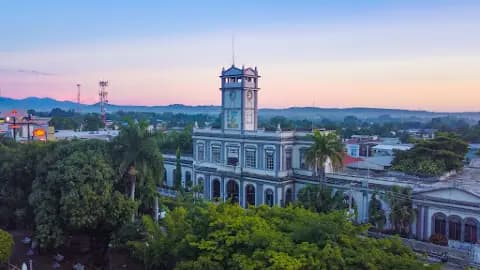 Aerial view of a historic clock tower building surrounded by lush green trees and distant hills during a colorful sunset in Santa Elena, Paraguay.