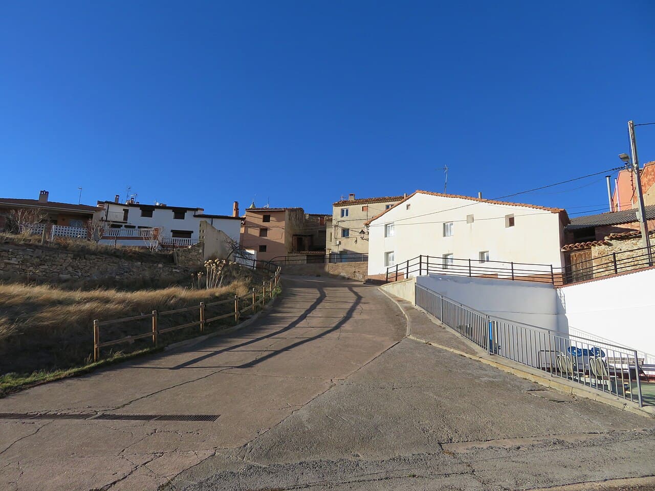 A sunlit, paved road winds uphill through a village with white and terracotta buildings under a clear blue sky, flanked by stone walls and fences.