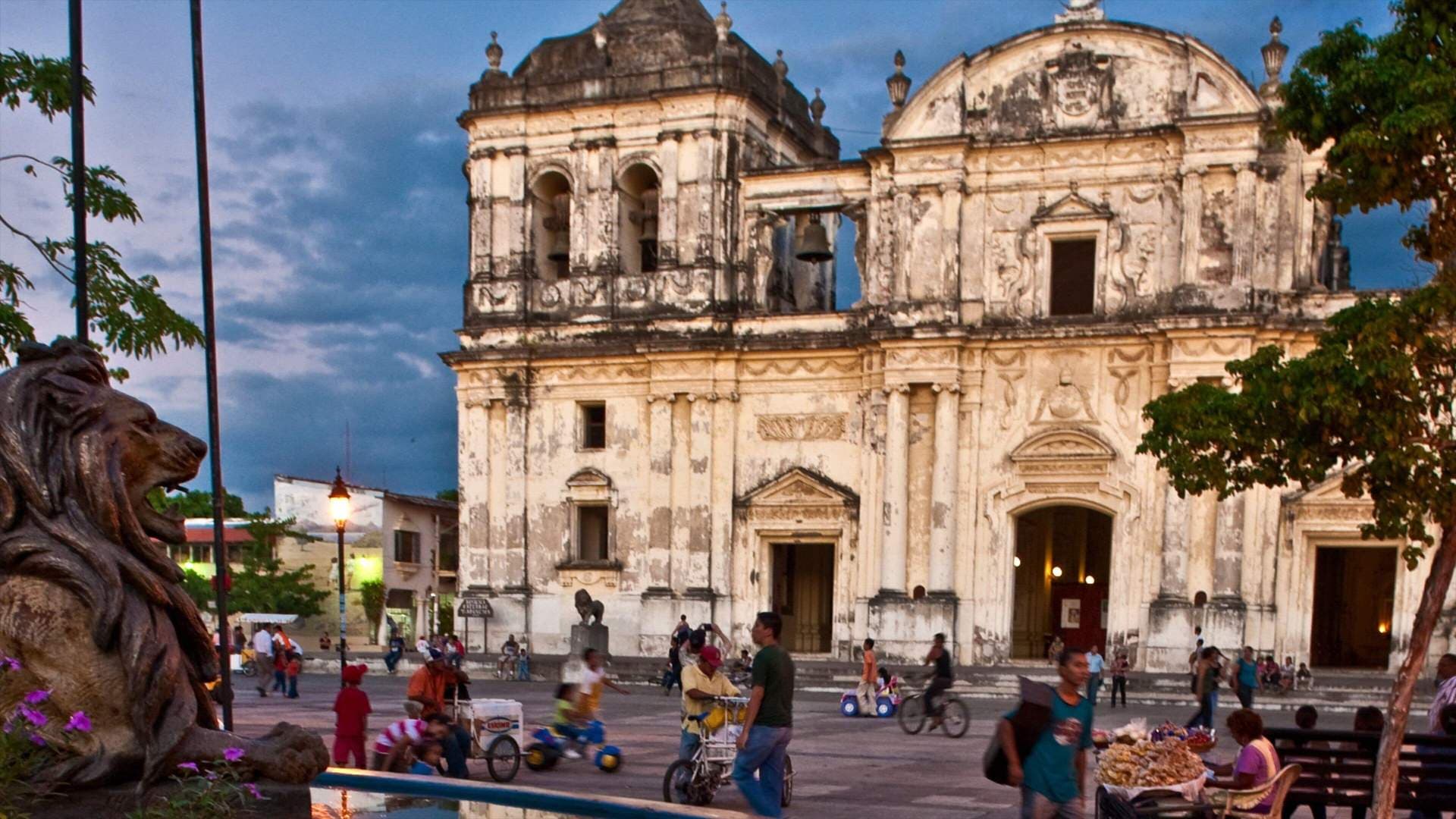 Wide dusk view of a colonial church facade and plaza with a lion statue, scattered pedestrians, and vendors.