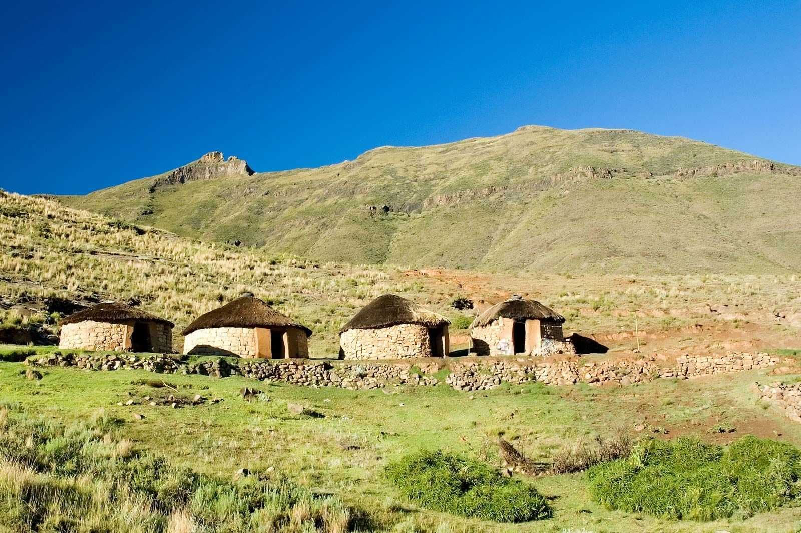 Four traditional stone and thatch huts sit on a grassy highland slope beneath rolling green mountains under a clear blue sky.