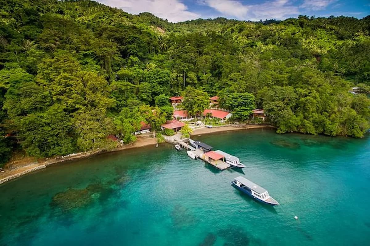 Aerial view of a tropical waterfront in Kabupaten Banggai, Indonesia, with a small dock, moored boats, and red-roofed buildings nestled among dense green hills