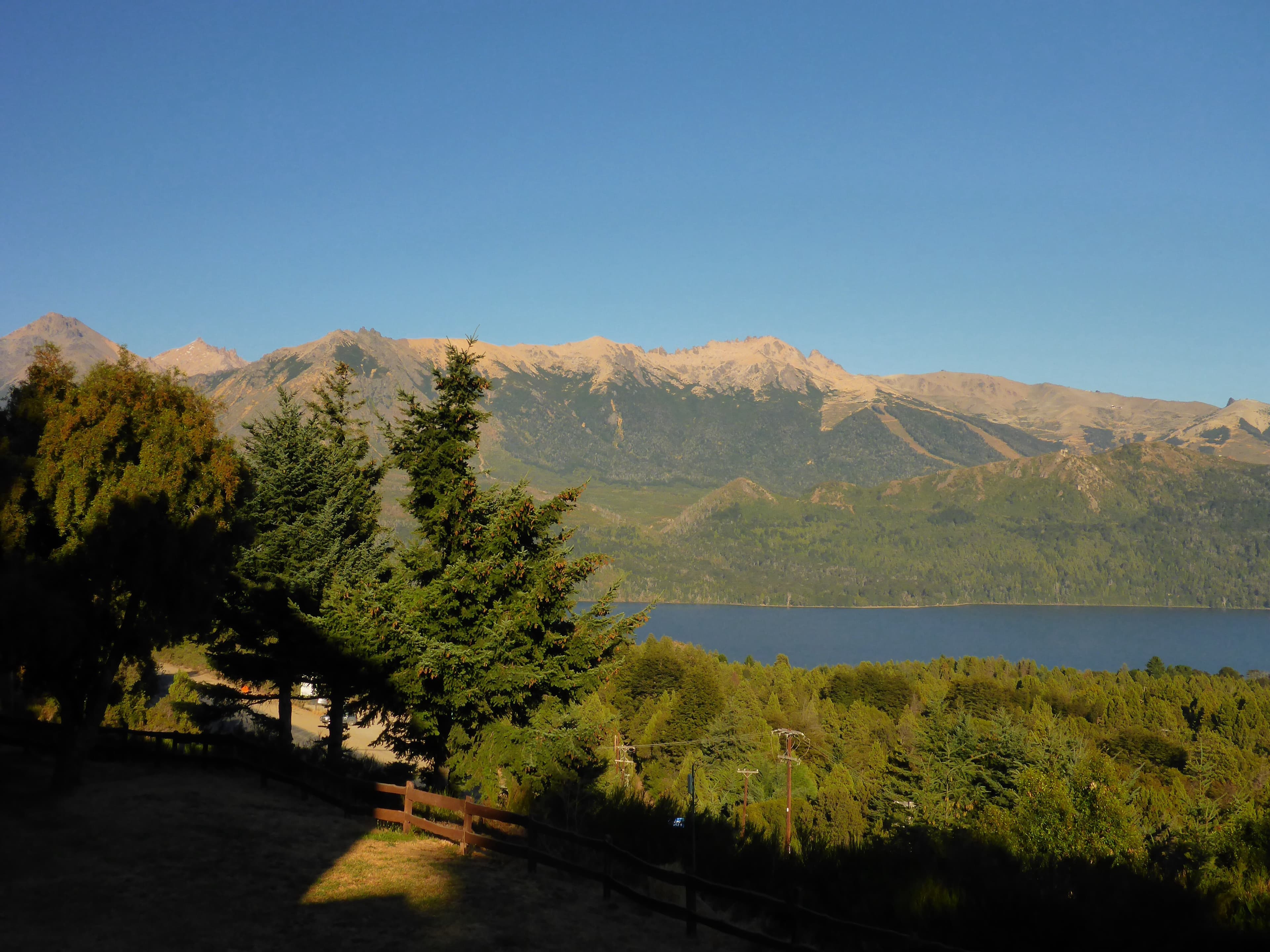 Scenic landscape featuring a blue lake and forested mountains under a clear sky, viewed from a grassy foreground with trees.