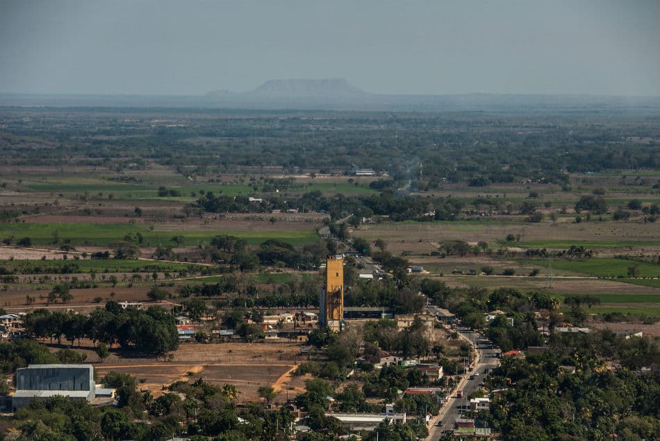 Aerial view of a rural landscape featuring a yellow industrial tower, scattered green fields, and a distant flat-topped mountain under a hazy sky.
