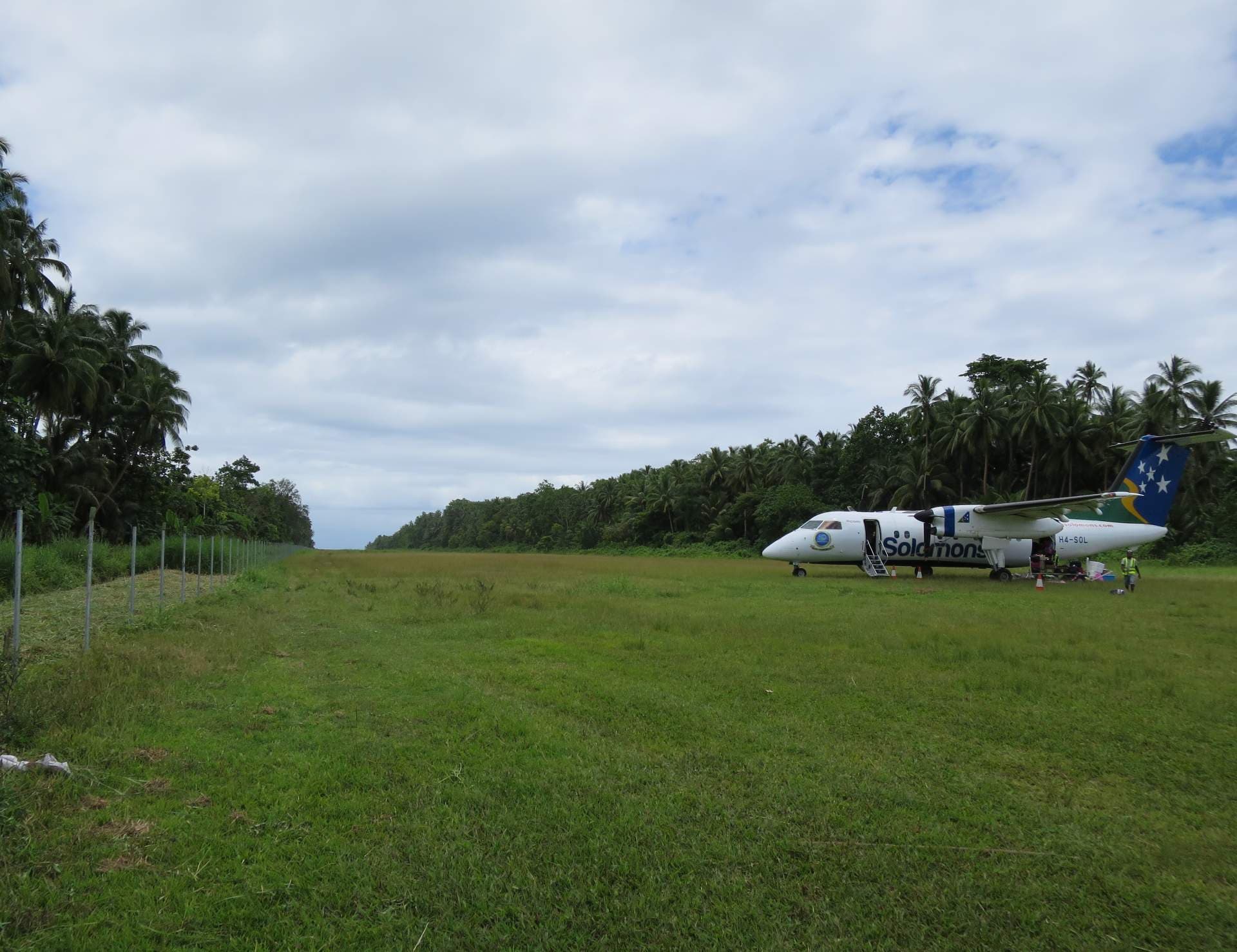 A Solomon Air turboprop aircraft parked on a grassy airstrip in Kirakira, Solomon Islands, surrounded by palm trees under a cloudy sky.