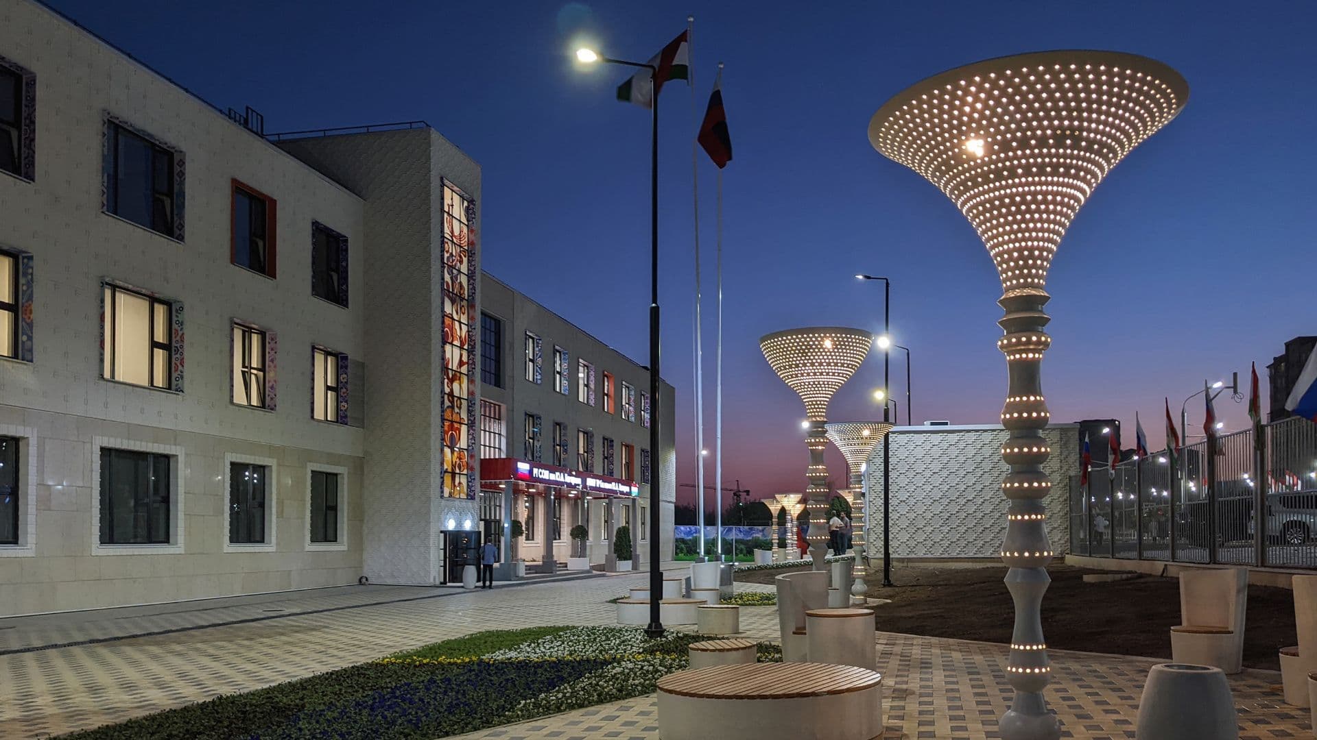 Evening street view of a modern plaza in Bokhtar featuring illuminated cone-shaped lamp posts, paved walkways, and contemporary buildings.