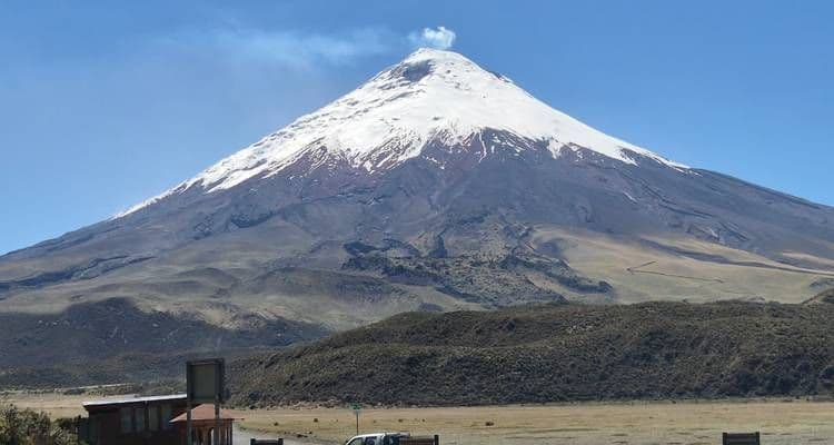 Snow-capped volcano under clear blue sky, viewed from highland terrain with small structures in the foreground near Atuntaqui, Imbabura.