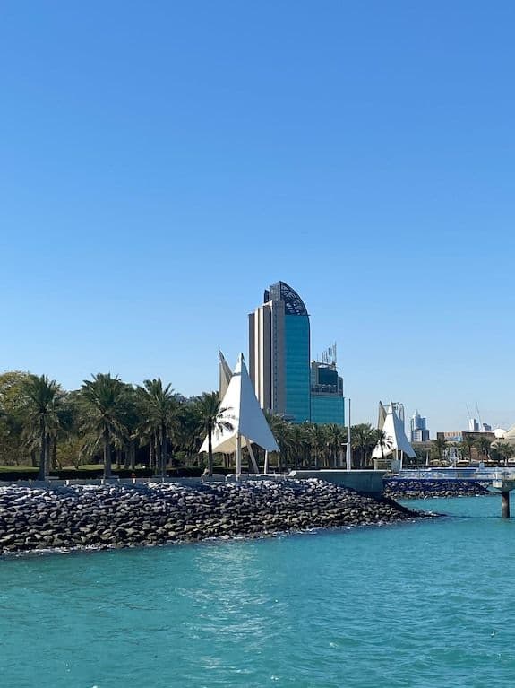 Wide waterfront view of the Salmiya promenade near Ash Shāmīyah with sail sculptures, palm trees, and a modern high-rise under a clear blue sky