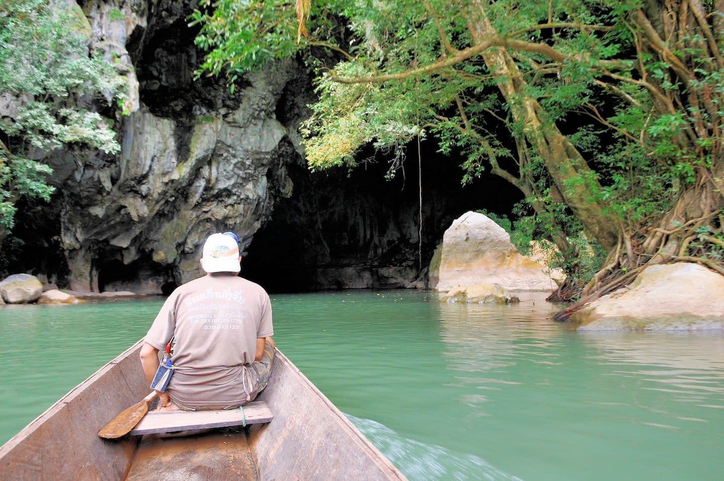 A boatman rows a wooden canoe on a turquoise river toward the dark, rocky entrance of Kong Lor Cave, surrounded by lush green vegetation.
