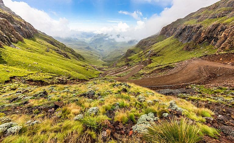 A winding dirt road descends through a green mountain valley with rocky slopes and distant clouds.
