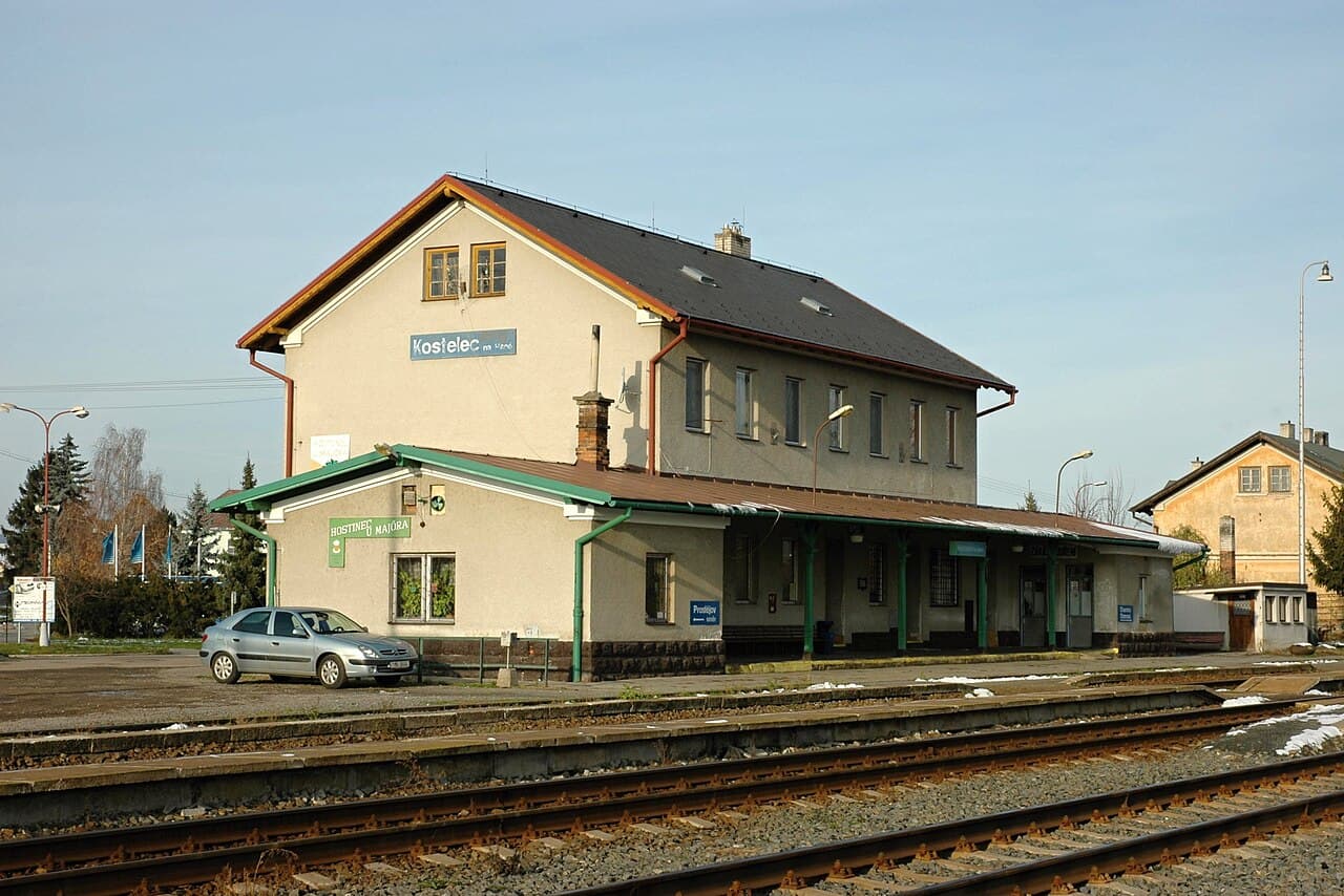 Exterior view of the Kostelec na Hané train station building, featuring tracks, a parked silver car, and clear signage under a blue sky.