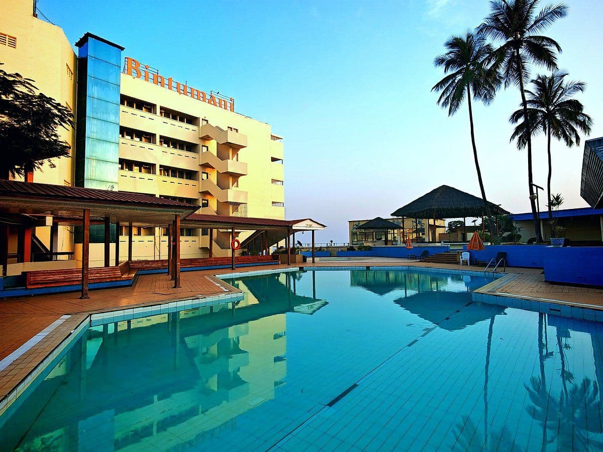 Exterior of Bintumani hotel with swimming pool, palm trees, and thatched-roof structures under a clear sky.
