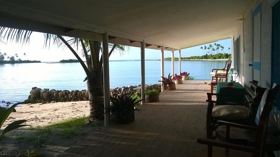 Covered wooden veranda of a seaside building overlooking calm water, sandy shore, palm trees, and the distant tropical coastline near Ambo Village, Kiribati.
