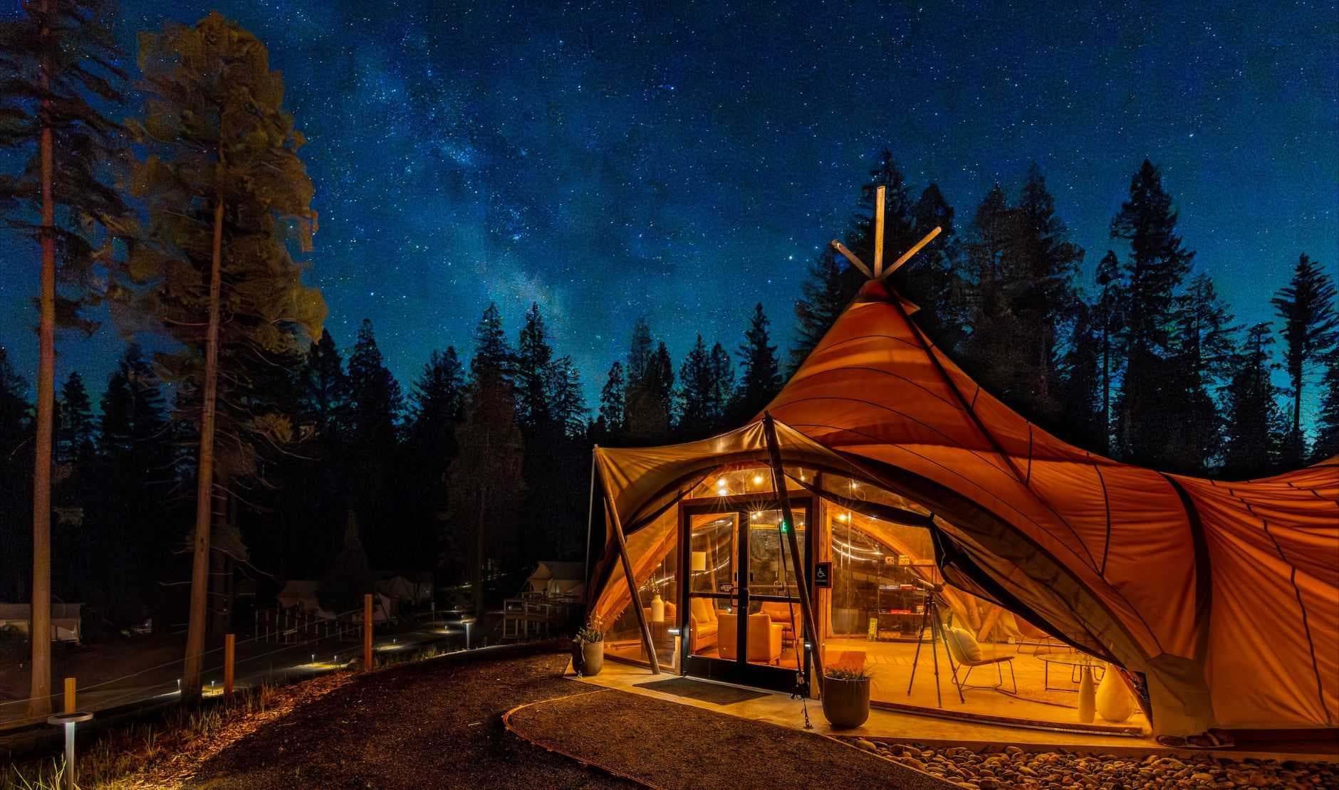 Illuminated geodesic glamping tent in a forest clearing at night, with a starry sky and surrounding trees.