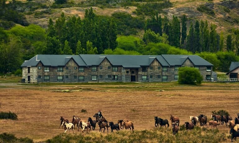 Wide exterior of a long, low stone and timber rural dwelling on open pampa with horses in the foreground.