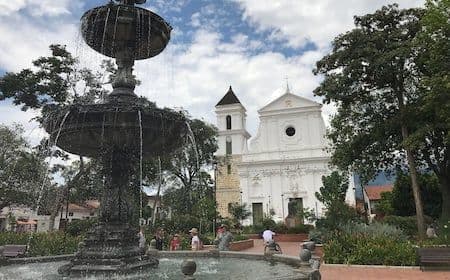 Wide plaza view in Liborina featuring a large fountain in the foreground and a white church with a bell tower in the background.