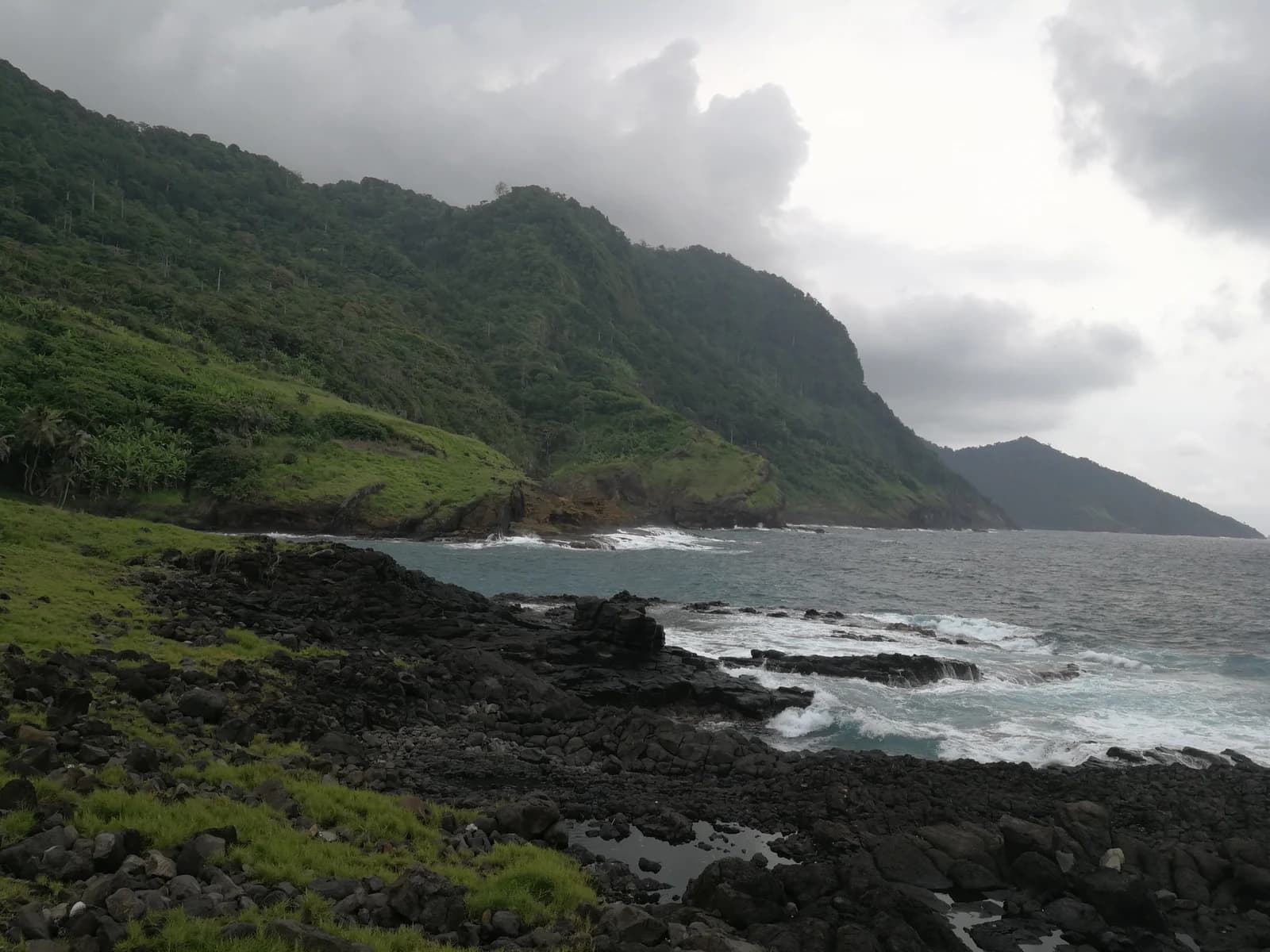 Coastal landscape featuring dark volcanic rocks, green hillsides, and ocean waves under an overcast sky.