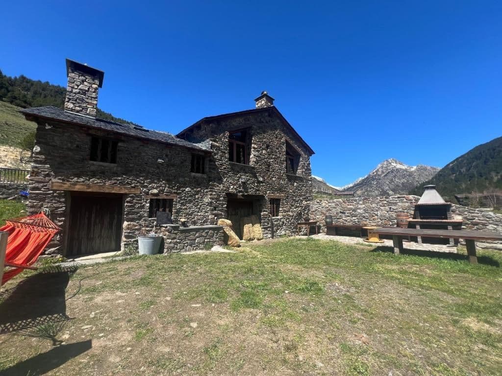 Rustic stone buildings in Canillo village under a clear blue sky, featuring a red hammock, wooden benches, and distant snow-capped mountains.