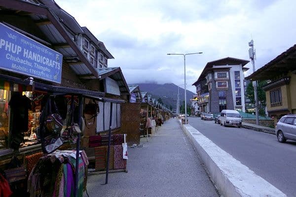 Street-level view of market stalls selling local goods along a road in Bhutan, with buildings and mountains in the background.