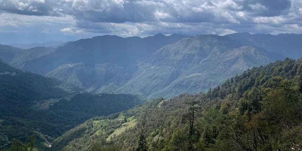 Wide landscape of a Bhutanese mountain valley with rolling forested hills and distant peaks.