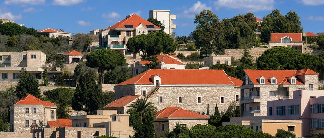 Wide exterior view of stone village buildings with red-tiled roofs on a hillside, including a church, under a blue sky.