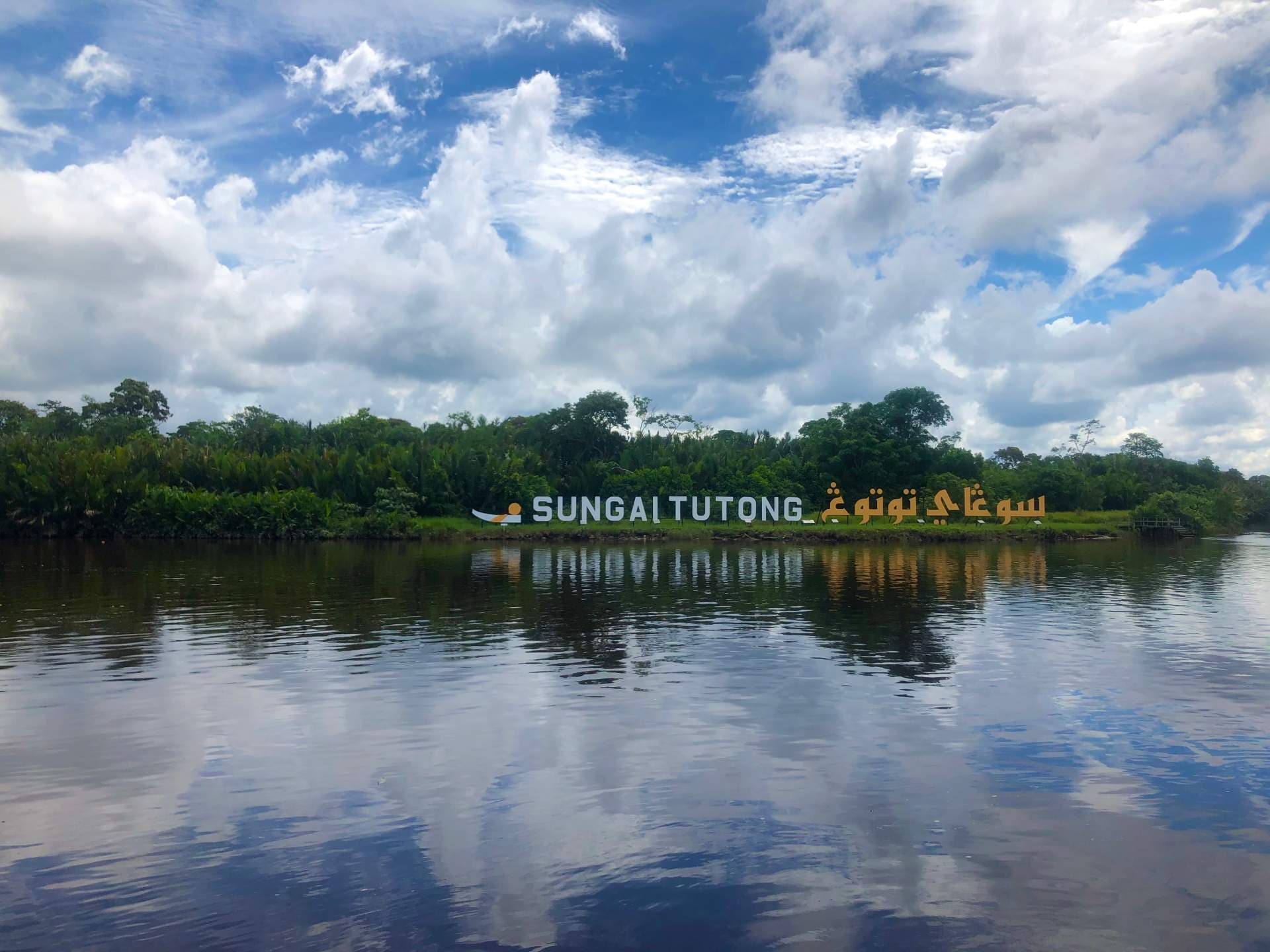 A scenic view of the Sungai Tutong river with a large sign reading 'SUNGAI TUTONG' on the lush green riverbank under a cloudy blue sky.
