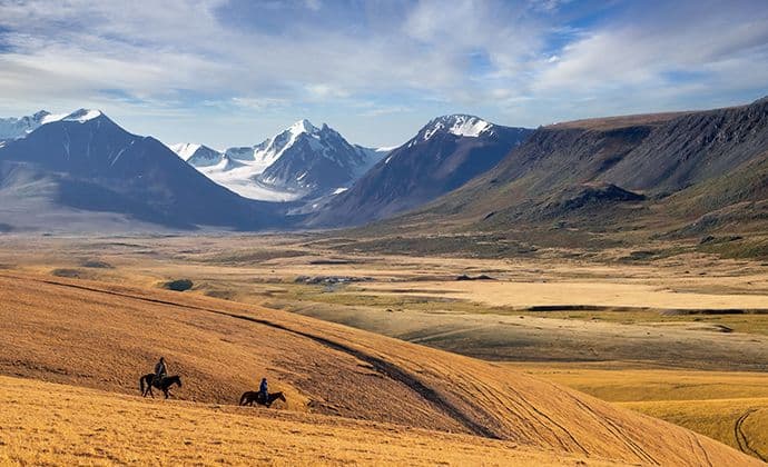 Wide landscape of golden autumn steppe hills with two distant riders, snow-capped mountains in the background under a partly cloudy sky.