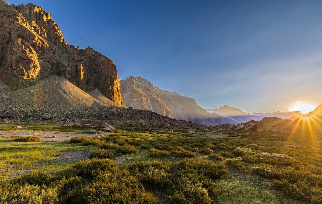 Mountain landscape at dusk with sun setting behind peaks, rocky slopes, and green shrubland foreground in the Copiapó Valley