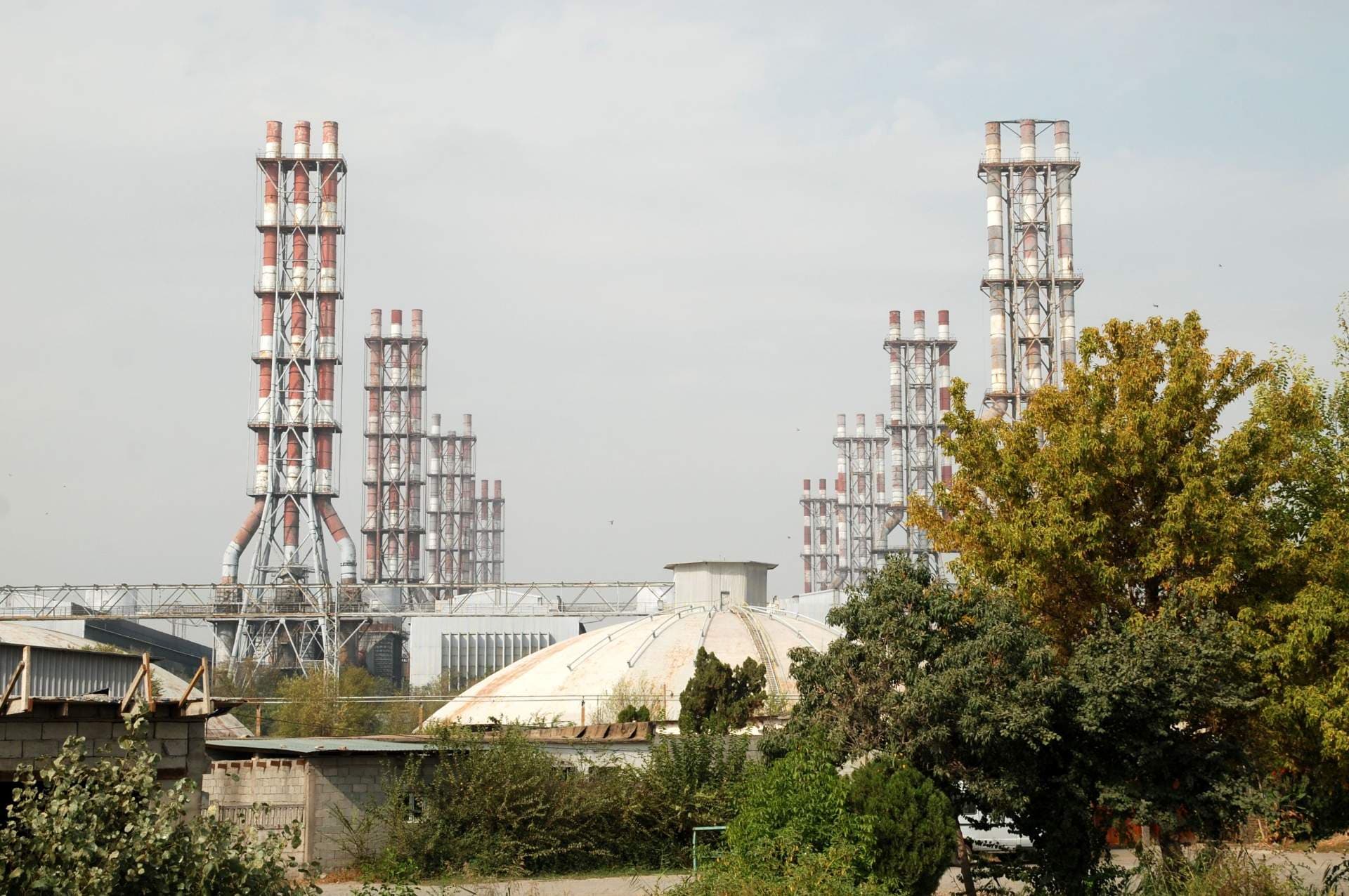 Wide exterior view of an industrial aluminum facility in Tursunzoda with tall red-and-white striped smokestacks, a domed structure, and surrounding greenery.