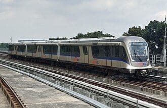 A commuter train travels on elevated tracks, with a station platform and trees visible in the background.