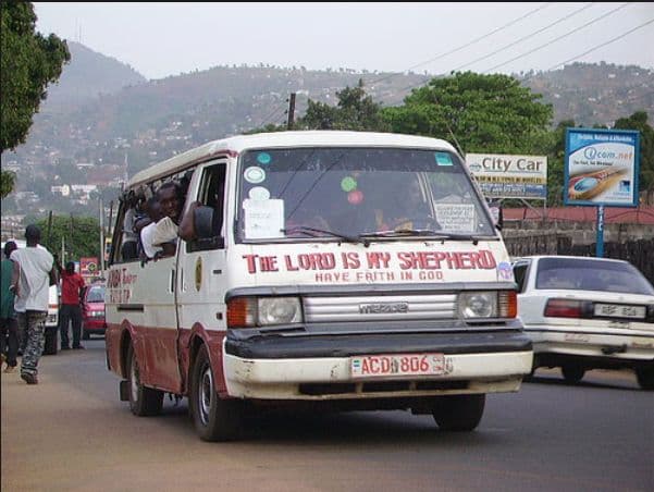 A white and red minibus with religious text on the front drives on a street, with pedestrians and hills in the background in Sierra Leone.
