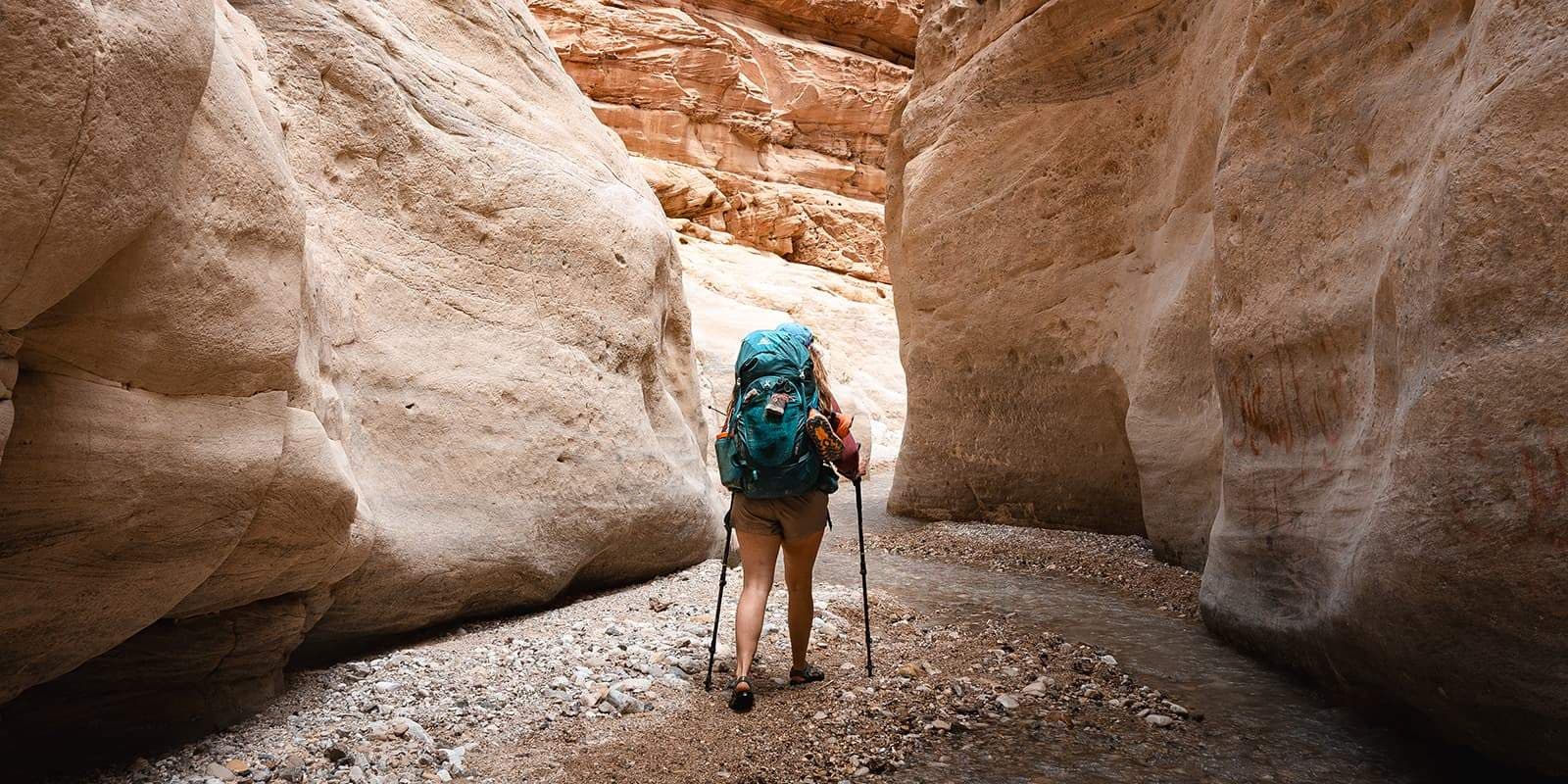 A hiker with a large blue backpack and trekking poles walks away from the camera along a rocky, unpaved trail through a narrow sandstone canyon.