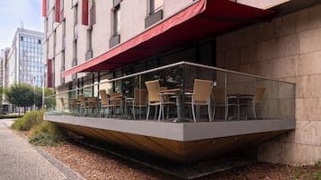 Street-level view of a modern building with a glass-enclosed terrace and red awning, situated on a paved sidewalk with urban architecture in the background.