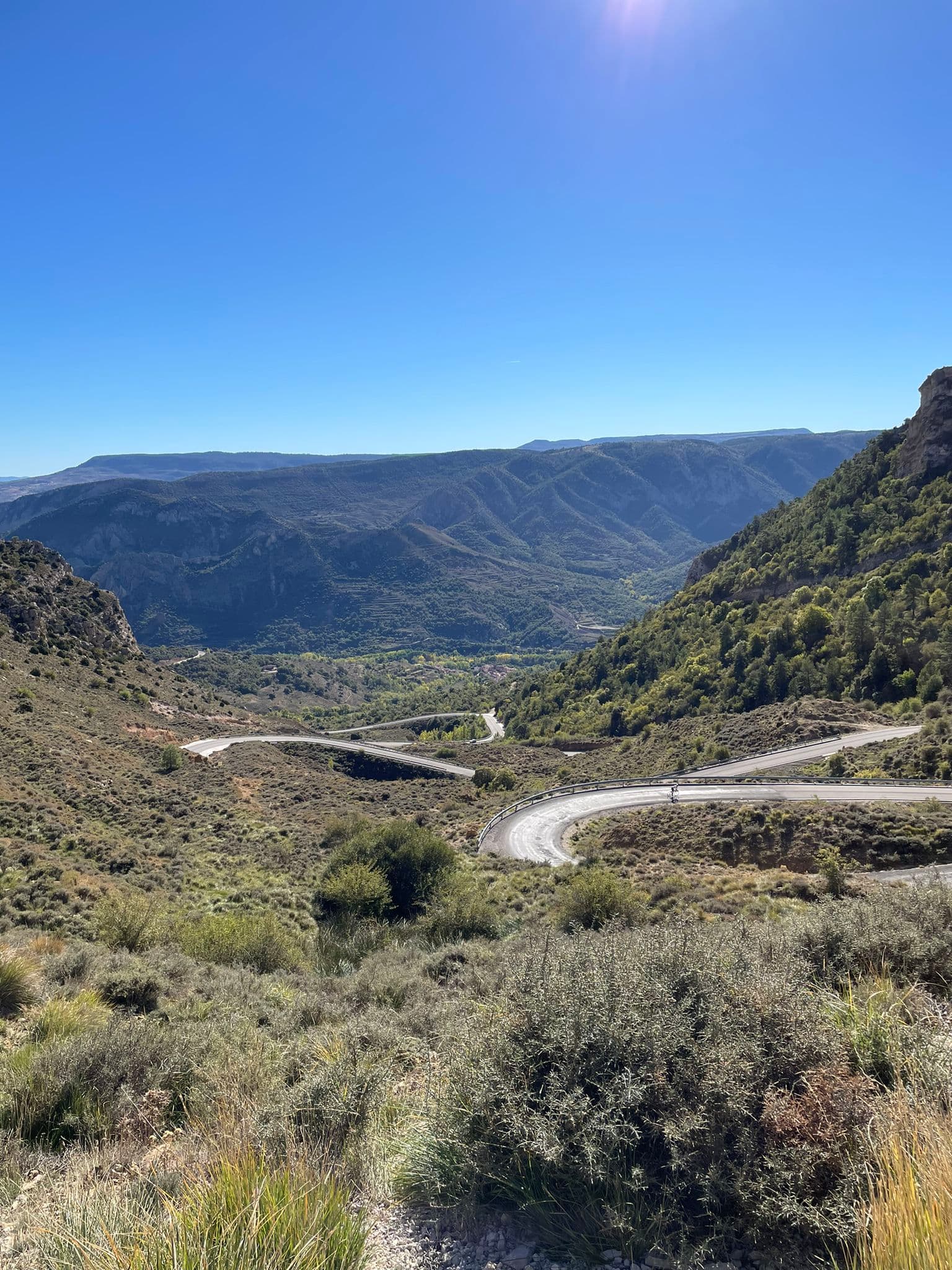 Winding mountain road with hairpin turns through a rugged, sunlit valley with green vegetation and distant hills under a clear blue sky.