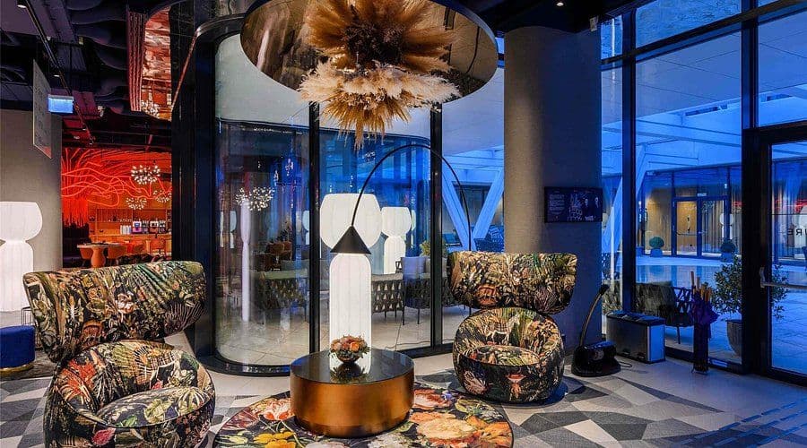 Modern lounge interior featuring floral patterned armchairs, a feather chandelier, and glass walls revealing blue-lit exterior.