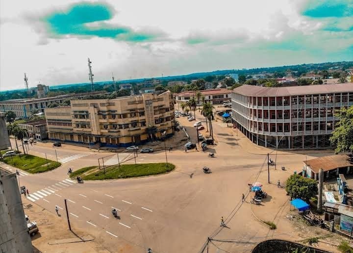 Aerial view of a wide intersection in Kisangani featuring multi-story buildings, a roundabout, and scattered traffic under a bright sky.