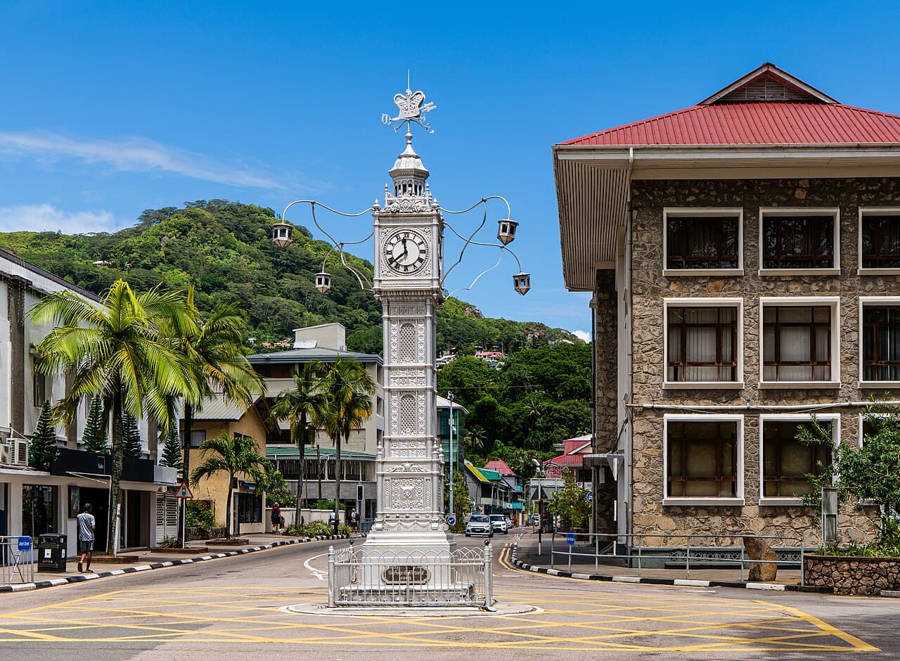 The Albert Clock stands prominently in a street intersection in Victoria, Seychelles, surrounded by stone buildings, palm trees, and lush green hills under a cl