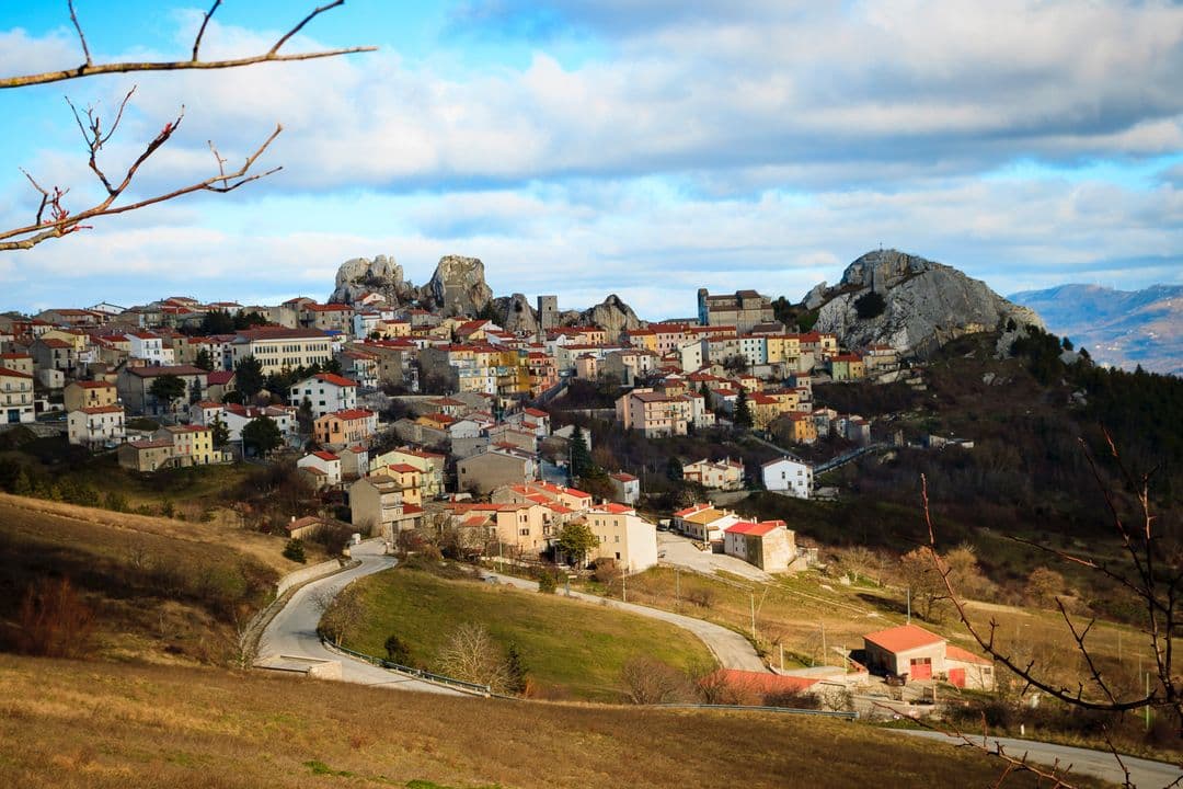 A hillside village with terracotta roofs nestled among rugged rocky peaks and dry grassy slopes under a cloudy sky.