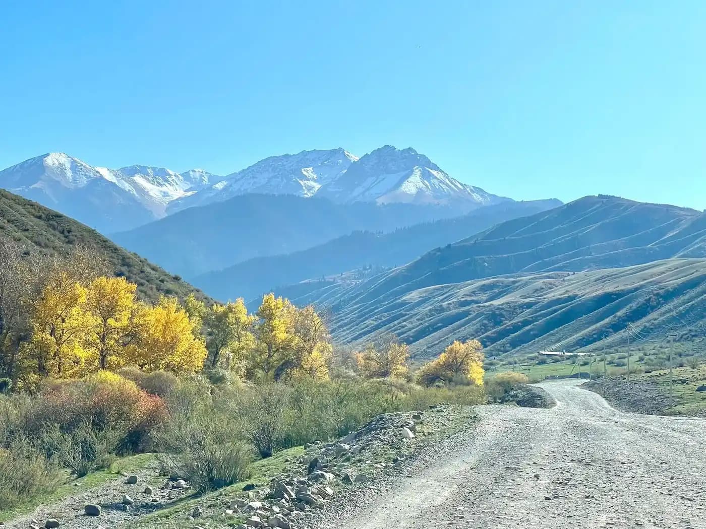 Unpaved gravel road winding through an autumn mountain valley with yellow foliage and distant snow-capped peaks near Bishkul, Kazakhstan.