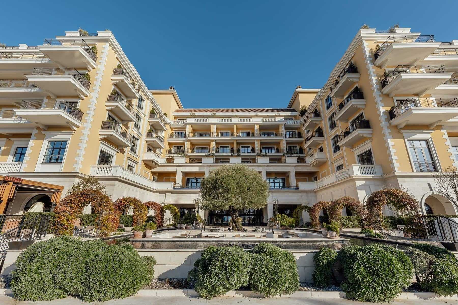 Yellow multi-story hotel exterior with white balconies, central courtyard, olive tree, and landscaped garden under clear blue sky.