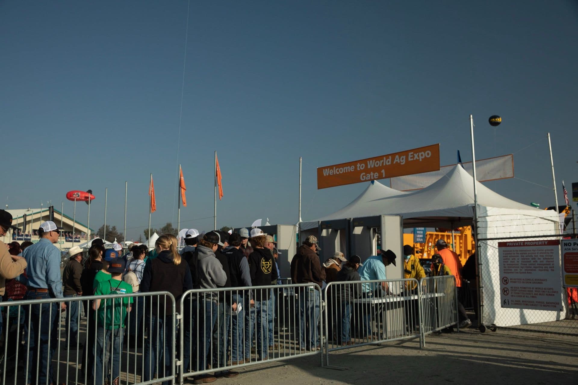 Crowd queues at World Ag Expo Gate 1 entrance with white tents, orange flags, and signage under clear sky.