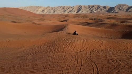 A lone vehicle traverses vast orange sand dunes with distant mountains, capturing the isolation of a desert landscape.